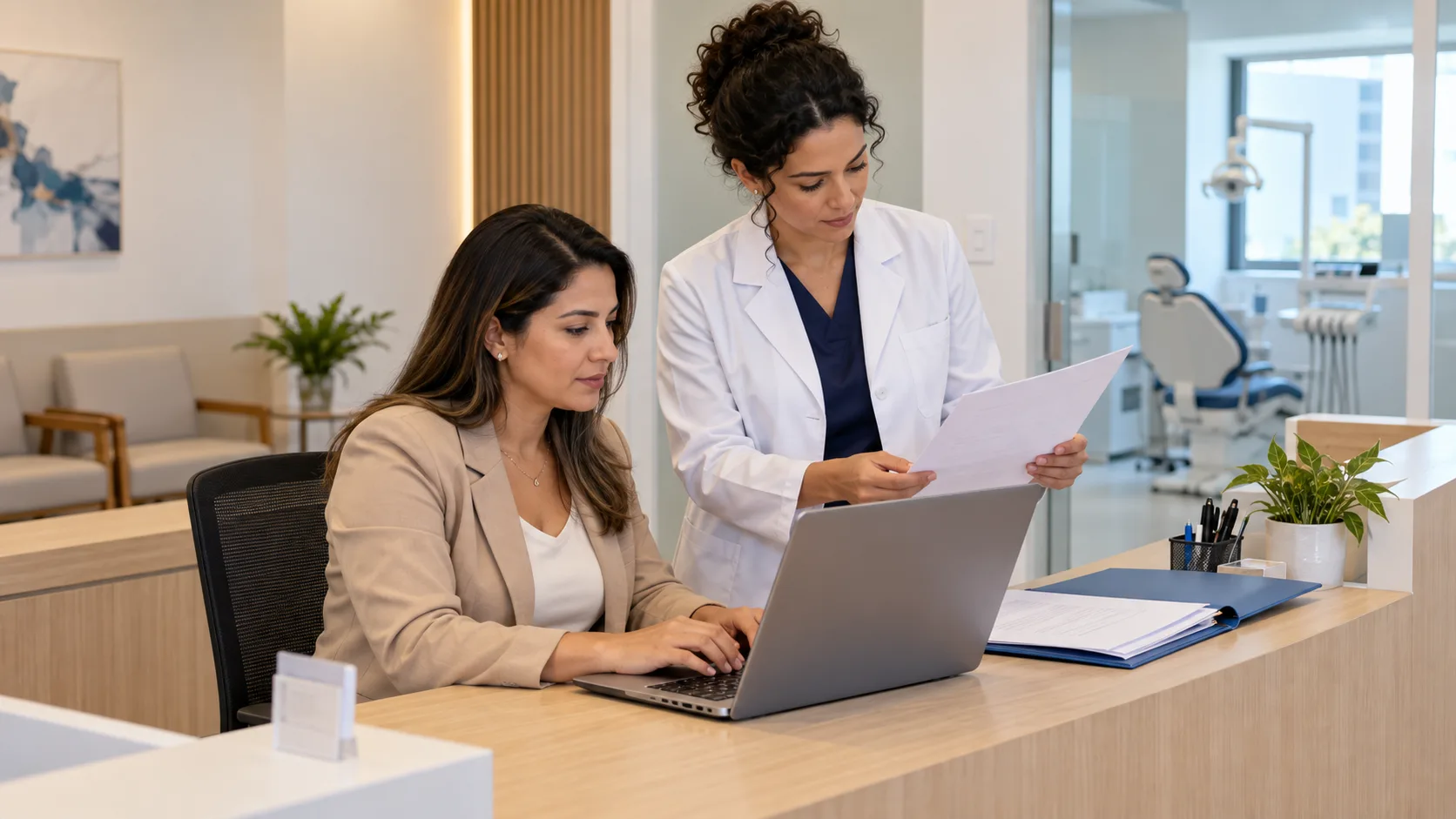 Gestora de clínica odontológica preenche um cadastro no notebook na recepção, com documentos da clínica numa pasta aberta ao lado, enquanto a dentista confere uma folha.